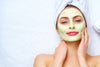 Woman with matcha face mask and towel on hair in front of a white background.